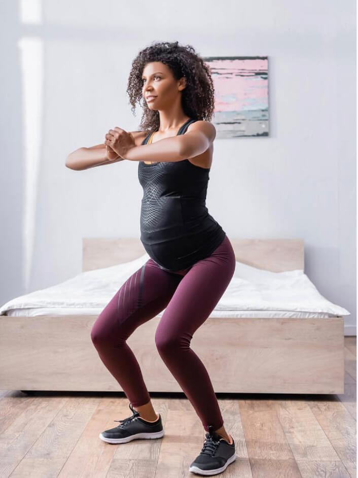 Pregnant black woman performing antenatal exercise squats in neutral bedroom with wooden floor wearing black and burgundy with running shoes
