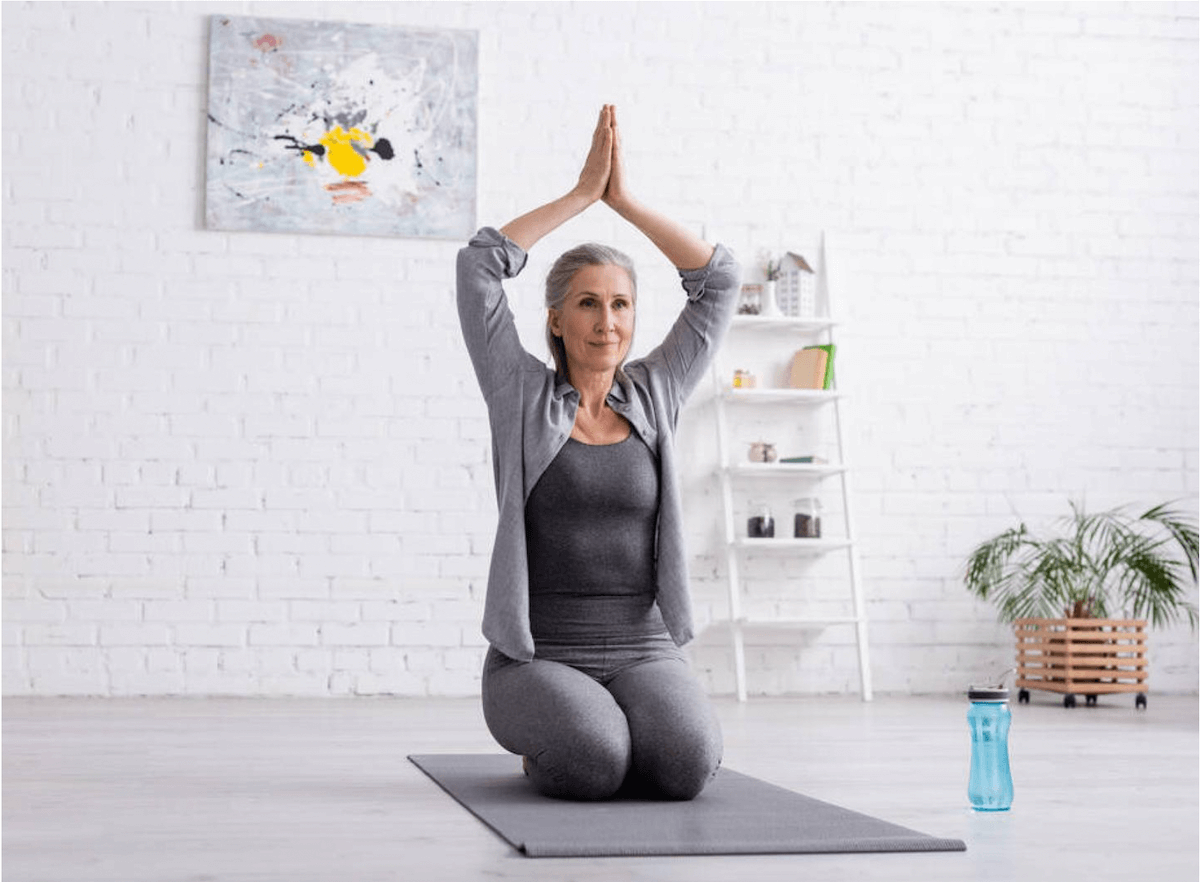 Older white woman with shoulder-length grey hair wearing grey workout clothes, performing yoga while sitting on a grey yoga mat in a white room with a brick wall behind her.  