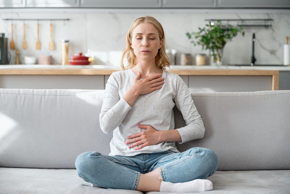 Blonde woman sitting on a sofa in front of a kitchen illustrating diaphragmatic breathing to support connecting the pelvic floor to the core.