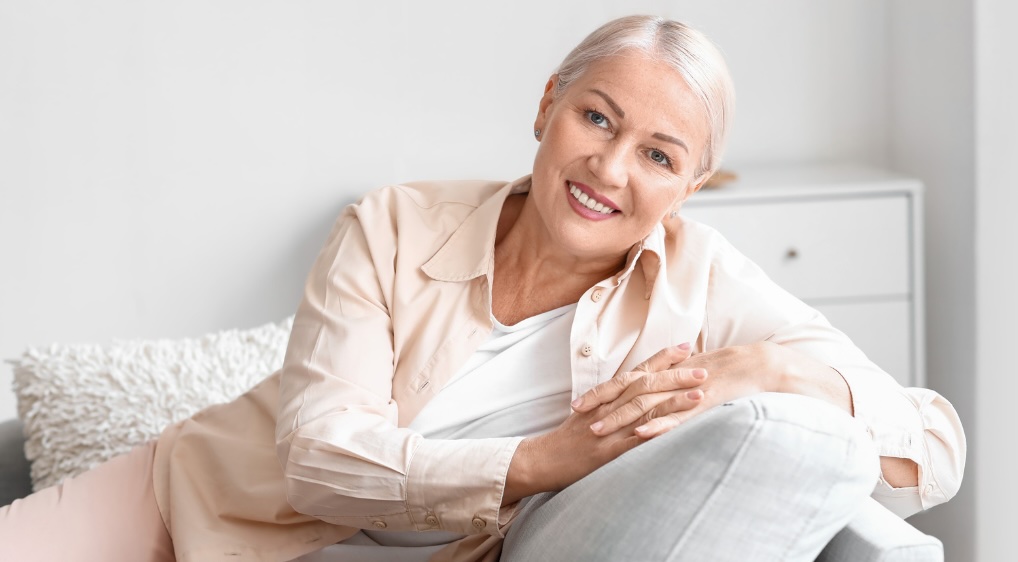 A middle-aged woman sitting on a sofa to illustrate the importance of health screenings for women in their 50s, 60s and 70s. 