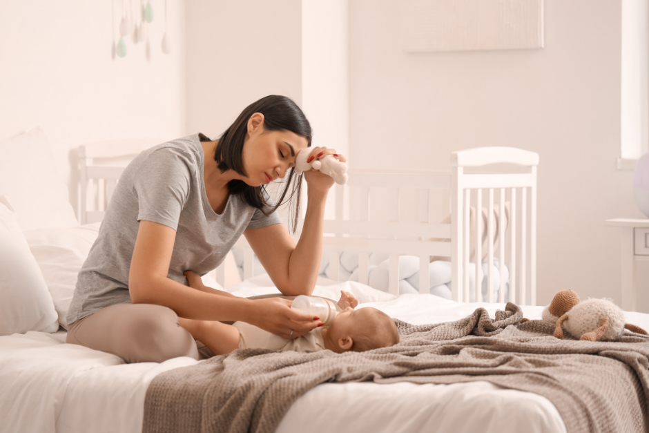 A young woman sitting on a bed feeding her baby with a bottle.