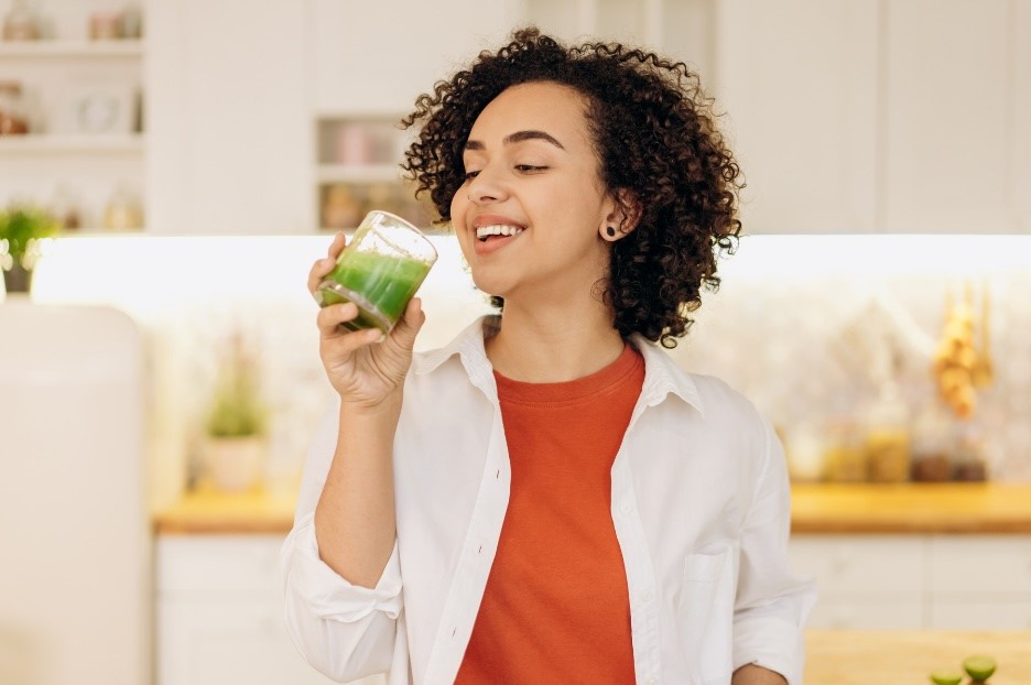 Woman standing in a kitchen wearing an orange tshirt and white shirt drinking a green juice to detox before pregnancy naturally.
