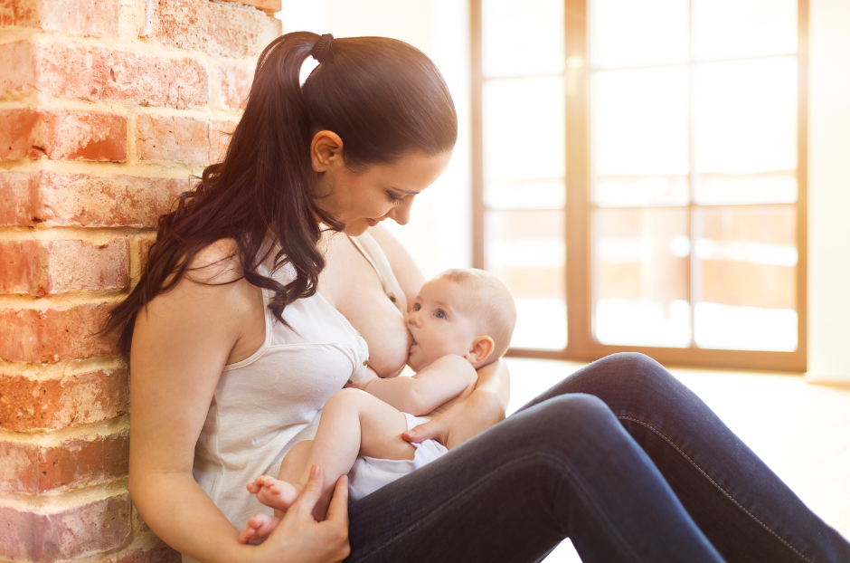 A smiling mum in a breastfeeding position sitting on the floor nursing a baby who is latched on. 