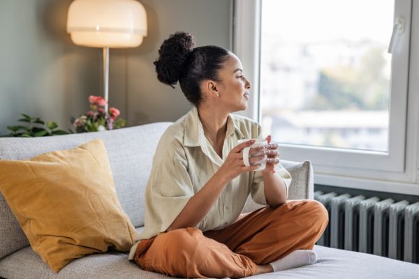 A young woman sitting on a sofa drinking tea to prioritise self-care and stress management.